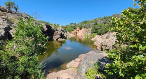 A serene natural landscape featuring a calm pond surrounded by rocky terrain and lush greenery under a clear blue sky.