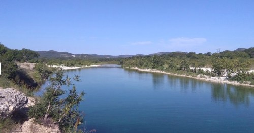 A serene river flows through lush greenery under a clear blue sky, surrounded by rolling hills in the distance.