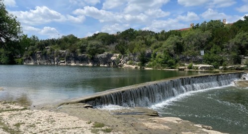 A serene landscape featuring a waterfall cascading over a stone ledge into a calm lake, surrounded by greenery and blue skies.
