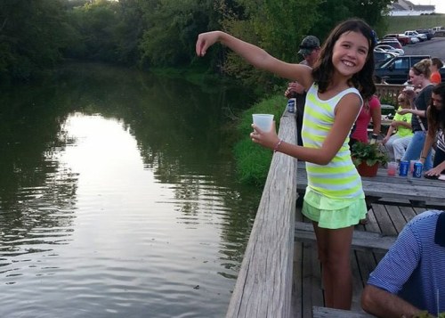 A girl in a striped shirt smiles and throws something into a calm river while others sit nearby on a wooden deck.