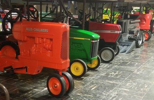A row of vintage toy tractors in various colors, including orange, red, and green, displayed on a dark floor.