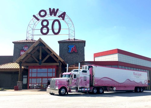 A pink truck parked outside the Iowa 80 truck stop, featuring a large sign and a clear blue sky.