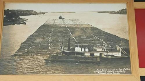 Historic black-and-white photo of a tugboat navigating a large log raft on a river.