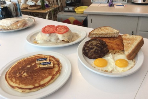 A table with three breakfast plates: pancakes with butter, a dish with eggs and sausage, and a plate of toast and hash browns.