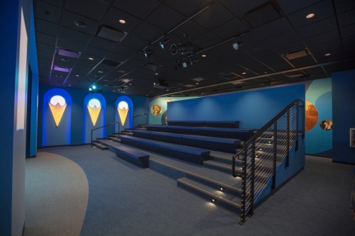 A modern auditorium with blue walls, tiered seating, and artistic wall features, illuminated by soft lighting.