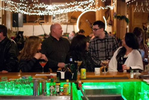 A lively bar scene with people chatting, drinks on the counter, and festive lights in a rustic setting.