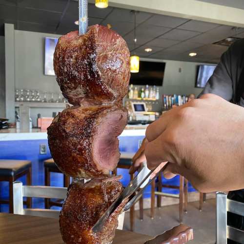 A hand using tongs to slice juicy, grilled meat from a skewer in a restaurant setting.