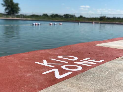 A red and gray concrete area by a water body, marked "KID ZONE," with colorful buoys floating in the water.