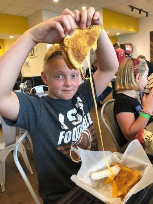 A boy holds up a grilled cheese sandwich, showcasing melted cheese stretching from the bread, smiling at the camera.