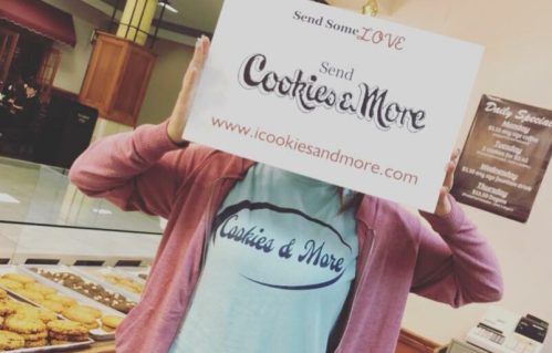 A person in a pink hoodie holds a sign for "Cookies & More" in a bakery, with cookies visible in the background.