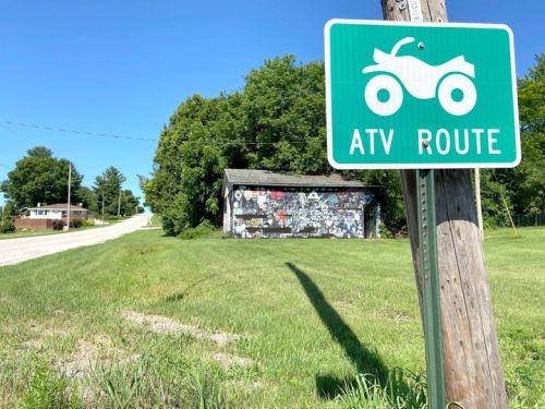 A green ATV route sign stands beside a grassy area, with a building covered in murals in the background.