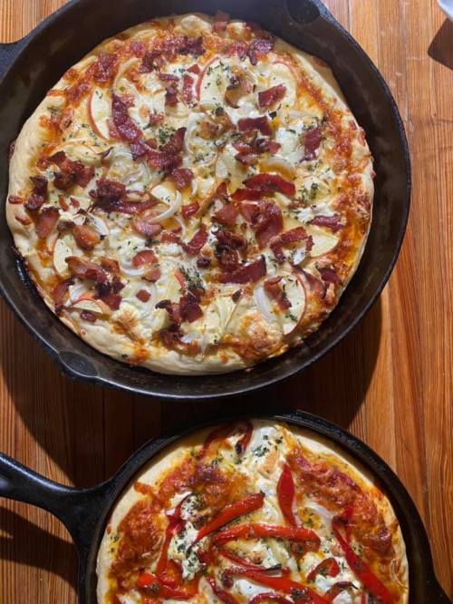 Two cast iron skillets with freshly baked pizzas: one topped with bacon and onions, the other with red peppers and cheese.