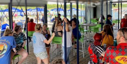 A lively gathering at a waterfront bar with people socializing, enjoying drinks, and seated at tables.