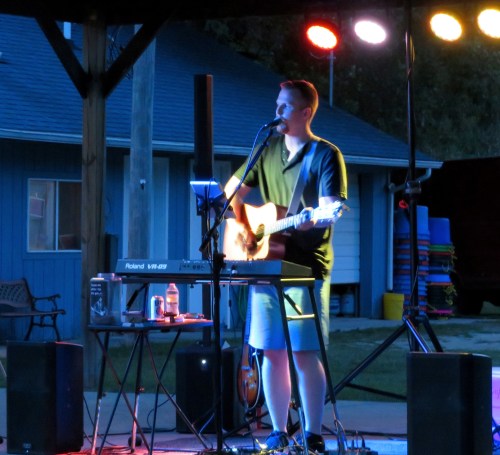 A musician performs on stage with a guitar and keyboard, illuminated by colorful lights in an outdoor setting.