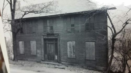 Black and white photo of an old, boarded-up wooden house surrounded by bare trees.