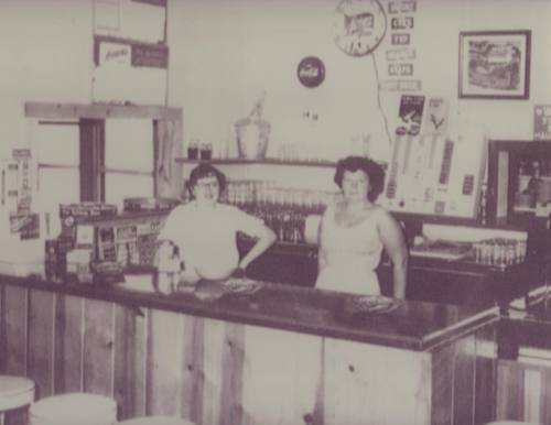 Two women stand behind a wooden counter in a vintage store, surrounded by soda bottles and old advertisements.