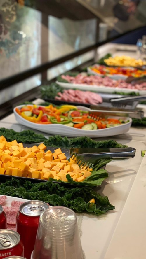 A buffet table with various salads, cheeses, and meats arranged on leafy greens, with drinks in the foreground.