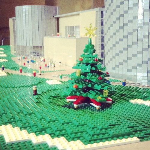 A LEGO model featuring a green Christmas tree with decorations, set on a grassy area near modern buildings.
