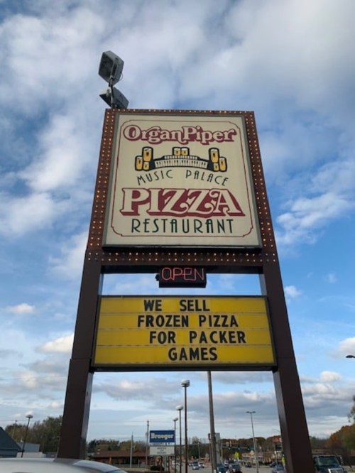 Sign for Organ Piper Music Palace Pizza Restaurant, advertising frozen pizza for Packer games. Open with a blue sky backdrop.