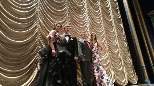 Four individuals in formal attire pose on stage in front of a draped curtain, smiling for the camera.