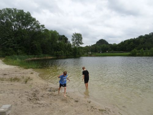 Two children play by a sandy shore, wading in a calm lake surrounded by trees under a cloudy sky.