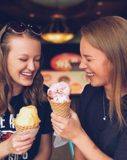 Two smiling friends enjoy ice cream cones, one with yellow and the other with pink ice cream, in a cheerful setting.