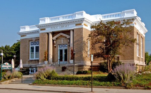 Historic public library building with columns, decorative facade, and landscaped entrance on a clear day.