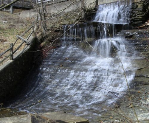 A serene waterfall cascading over rocky terrain, surrounded by bare trees and a stone wall.