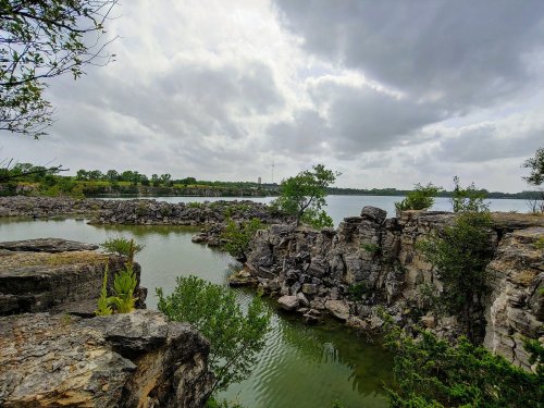 A serene landscape featuring rocky outcrops and calm water under a cloudy sky, surrounded by greenery.