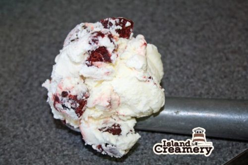 A scoop of red velvet cake ice cream with chunks, resting on a metal scoop against a gray background.
