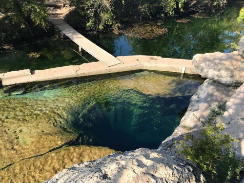 A clear blue swimming hole surrounded by rocks and trees, with a wooden bridge crossing over the water.