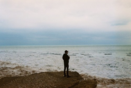 A person stands on a rocky shore, gazing out at a calm, icy ocean under a cloudy sky.