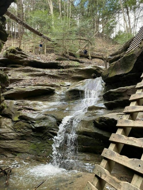 A small waterfall cascades over rocks in a wooded area, with trees and a wooden structure in the background.