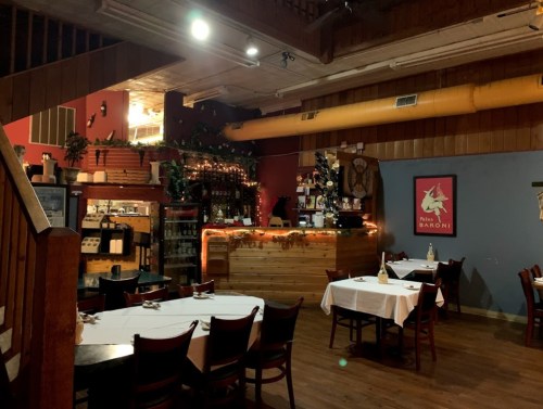 Cozy restaurant interior with wooden decor, tables set for dining, and a warmly lit bar area in the background.