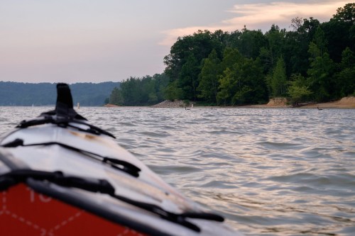 A kayak on calm water, with a lush green shoreline and trees in the background under a soft, cloudy sky.