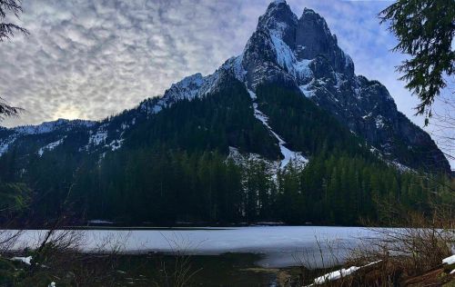 A majestic mountain with snow-capped peaks rises above a frozen lake, surrounded by evergreen trees and a cloudy sky.