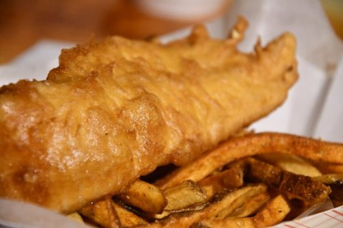 A close-up of a golden-brown battered fish fillet served with crispy fries on a paper tray.