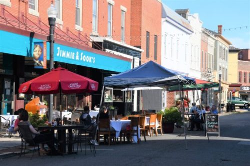 Outdoor dining scene with tables under umbrellas, featuring a restaurant and a street lined with historic buildings.