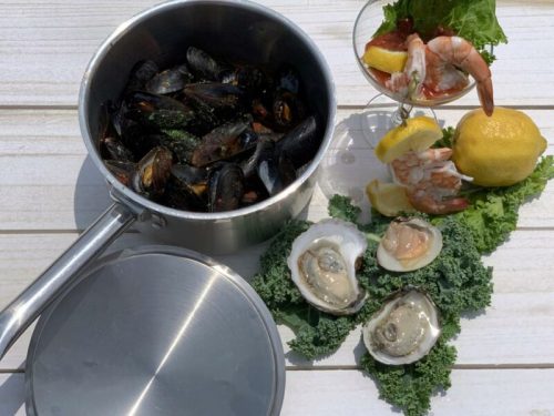 A pot of mussels beside a seafood platter with shrimp, oysters, lemon, and lettuce on a wooden surface.