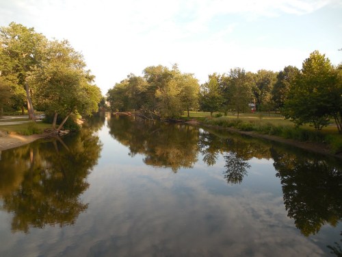 A calm river reflects trees and a clear sky, surrounded by a park with green grass and walking paths.