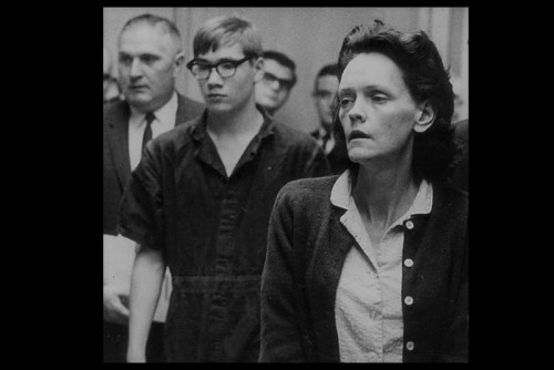 A black and white photo of two individuals in a courtroom, one in a jumpsuit and the other in a cardigan, looking serious.