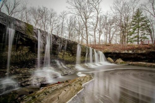 A serene waterfall cascades over rocky ledges into a calm pool, surrounded by bare trees and a misty atmosphere.