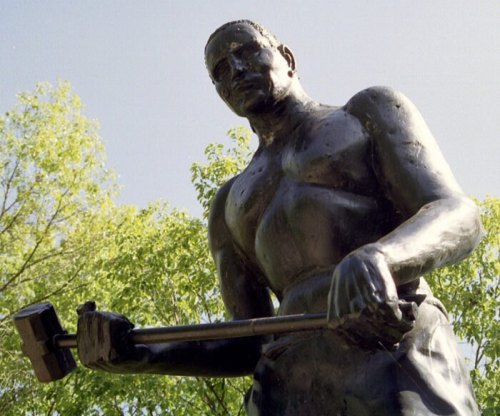 A bronze statue of a muscular man holding a hammer, surrounded by green trees under a clear sky.