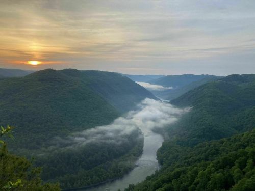 A serene landscape of rolling green hills and a winding river, with mist rising and a soft sunset in the background.