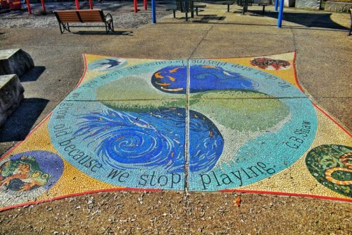 Colorful mosaic on a playground floor featuring waves, animals, and the quote, "We don't stop playing because we grow old."