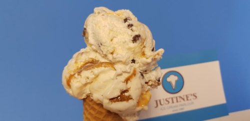 A close-up of a creamy ice cream cone with swirls of caramel and chocolate chips, next to a Justine's Ice Cream Parlour sign.