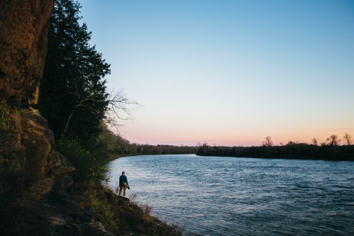 A person stands by a riverbank at sunset, surrounded by trees and rocky cliffs, with a calm river flowing.