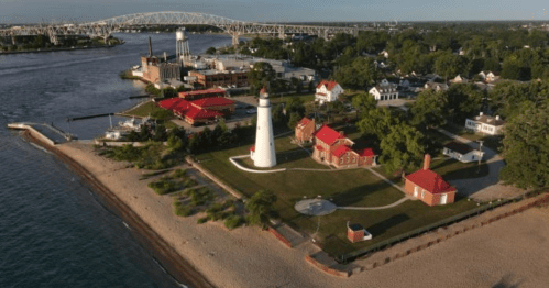 Aerial view of a lighthouse surrounded by red-roofed buildings, sandy beach, and a river with a bridge in the background.