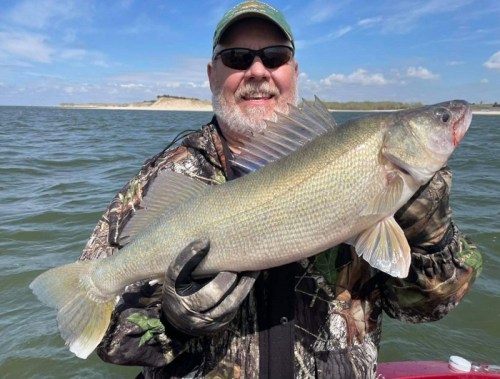 A smiling man in camouflage holds a large walleye fish, standing on a boat with a scenic water background.