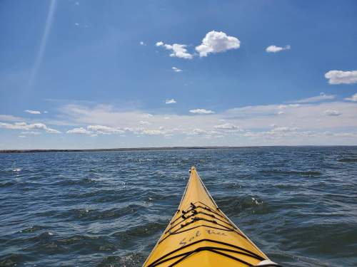 A yellow kayak glides over blue water under a bright sky with scattered clouds.